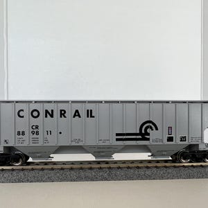 Può includere: Un vagone ferroviario modello CONRAIL grigio con ruote nere e un design dettagliato. Il vagone presenta il logo CONRAIL e vari contrassegni, tra cui "CR 88 98 11". È posizionato su un binario modello.