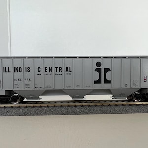Può includere: Un modello di carro merci Illinois Central grigio con scritte e logo neri. Sono visibili le parole "ILLINOIS CENTRAL" e "MAIN LINE OF MID-AMERICA". Il modello è su un binario.