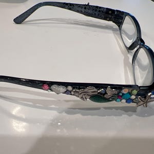 Könnte beinhalten: Dunkelblaue Brille mit dekorativen Verzierungen. Die Bügel zeigen ein Meerjungfrau-, Seestern- und Herzdesign mit bunten Edelsteinen. Die Fassungen haben ein Blumenmuster in der Nähe der Scharniere.