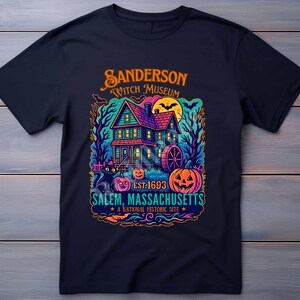 May include: Navy blue t-shirt featuring a colorful graphic of the Sanderson Witch Museum in Salem, Massachusetts. The design includes a house, pumpkins, and the text "EST. 1693". A national historic site.