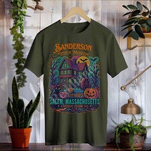 May include: Olive green t-shirt featuring a colorful graphic of a haunted house with the text "Sanderson Witch Museum" and "Salem, Massachusetts". The design includes pumpkins, a black cat, and a full moon, with the text "Est. 1693".