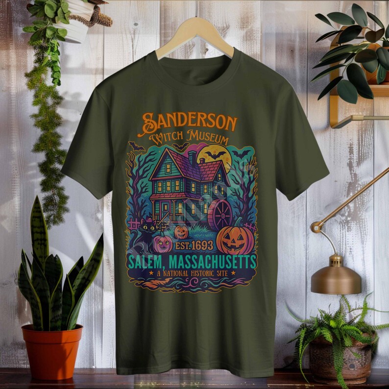 May include: Olive green t-shirt featuring a colorful graphic of a haunted house with the text "Sanderson Witch Museum" and "Salem, Massachusetts". The design includes pumpkins, a black cat, and a full moon, with the text "Est. 1693".