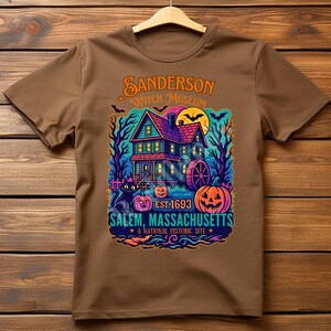 May include: Brown t-shirt featuring a colorful graphic of a haunted house with pumpkins, bats, and the text "Sanderson Witch Museum, Salem, Massachusetts, Est. 1693." The design is in shades of blue, orange, purple, and green.