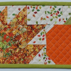 May include: A quilted fabric placemat with an autumn theme. The placemat features a large orange pumpkin with a green stem and a patchwork of autumn leaves in shades of brown, orange, yellow, and green.
