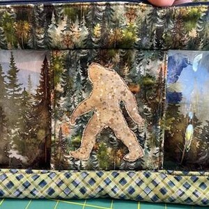 May include: A zippered bag with a forest-themed design. The bag features a Bigfoot applique in the center, with forest scenes on either side. The top and bottom have a forest print and a plaid pattern, respectively. The bag is designed for storage.