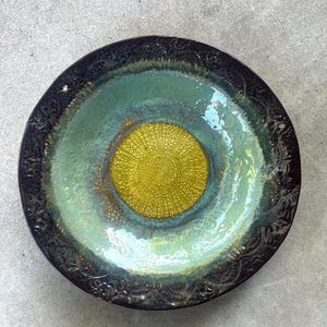 May include: A round ceramic bowl featuring a dark brown, embossed rim with intricate patterns. The center displays a vibrant, textured yellow circular design, surrounded by a smooth, glossy transition of light green and teal glaze.