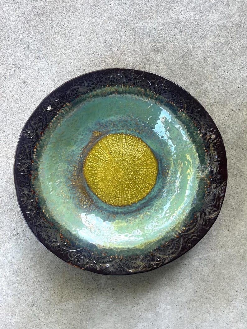 May include: A round ceramic bowl featuring a dark brown, embossed rim with intricate patterns. The center displays a vibrant, textured yellow circular design, surrounded by a smooth, glossy transition of light green and teal glaze.