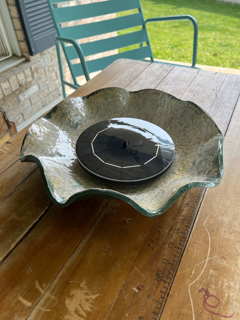 May include: A black solar powered water fountain sits in a green and white wavy ceramic bowl. The fountain is on a wooden table.