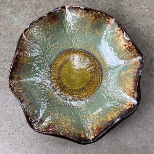 May include: A ceramic bowl with an organic, wavy edge. The bowl has a textured floral pattern in green and gold, with a dark brown rim. The centre has a circular design. A decorative piece, ideal for serving or display, with a diameter of approximately 30 cm.