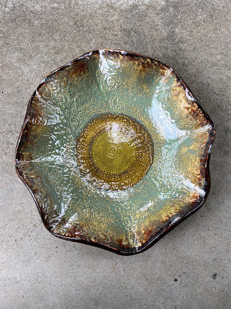 May include: A large, green and brown ceramic bowl with a decorative, embossed design. The bowl has a scalloped edge and a slightly raised centre.