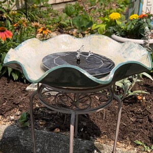 May include: A white and green ceramic bird bath with a solar powered fountain. The bird bath is on a metal stand with a decorative design. The bird bath is surrounded by green plants and brown mulch.
