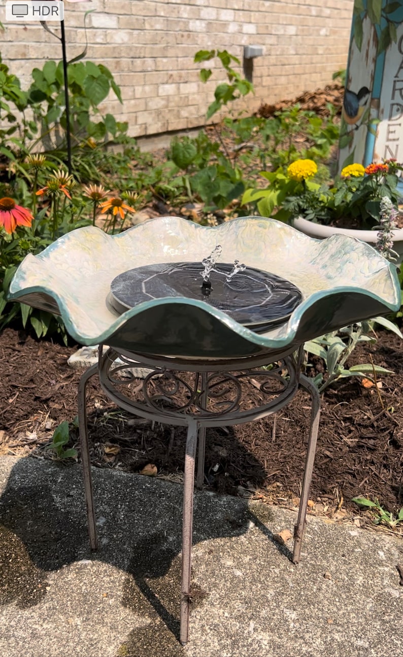 May include: A white and green ceramic bird bath with a solar powered fountain. The bird bath is on a metal stand with a decorative design. The bird bath is surrounded by green plants and brown mulch.