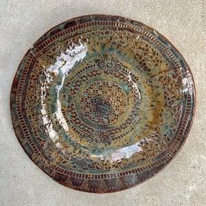 May include: A round ceramic plate featuring a glossy glaze in shades of brown, bronze, and teal. The surface is decorated with intricate, embossed lace-like patterns, including floral motifs and geometric borders that cover the entire plate.