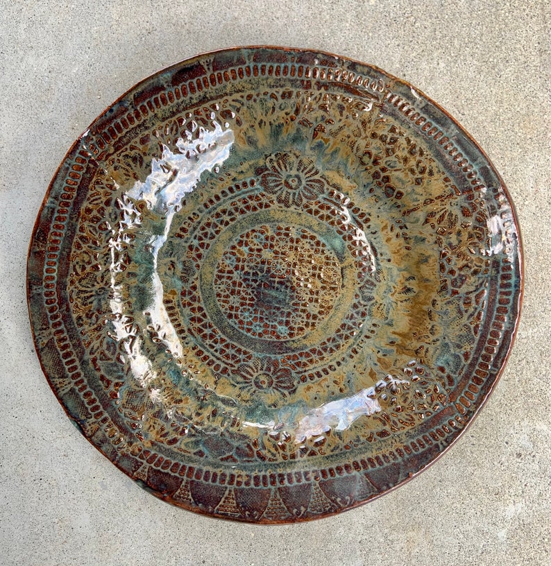 May include: A round ceramic plate featuring a glossy glaze in shades of brown, bronze, and teal. The surface is decorated with intricate, embossed lace-like patterns, including floral motifs and geometric borders that cover the entire plate.