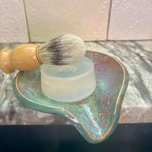 Op de afbeelding: Een witte scheerschuimzeep en een houten scheerkwast met een zwart-wit borstelkop liggen op een teal en gouden keramische schaal.