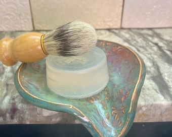 May include: A white shaving soap puck and a wooden shaving brush with a black and white bristle head are sitting on a teal and gold ceramic dish.