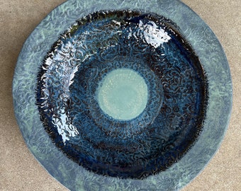 May include: A handmade ceramic plate with a unique design. The plate features a central light blue circle, surrounded by concentric rings of dark blue and black patterns. The outer rim is a lighter blue with a floral pattern.