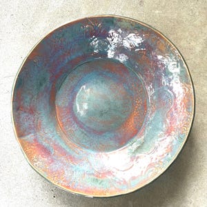 May include: A round ceramic bowl featuring a glossy, multi-colored glaze with swirling patterns of teal, blue, and copper-orange. The surface displays an intricate, embossed floral or scrollwork texture throughout, with a smooth, reflective finish.