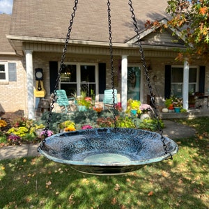 May include: A round, shallow ceramic bird bath with a textured, dark blue and black glaze featuring intricate, swirling patterns. The basin is suspended by three black metal chains attached to the rim, creating a hanging structure for outdoor use.