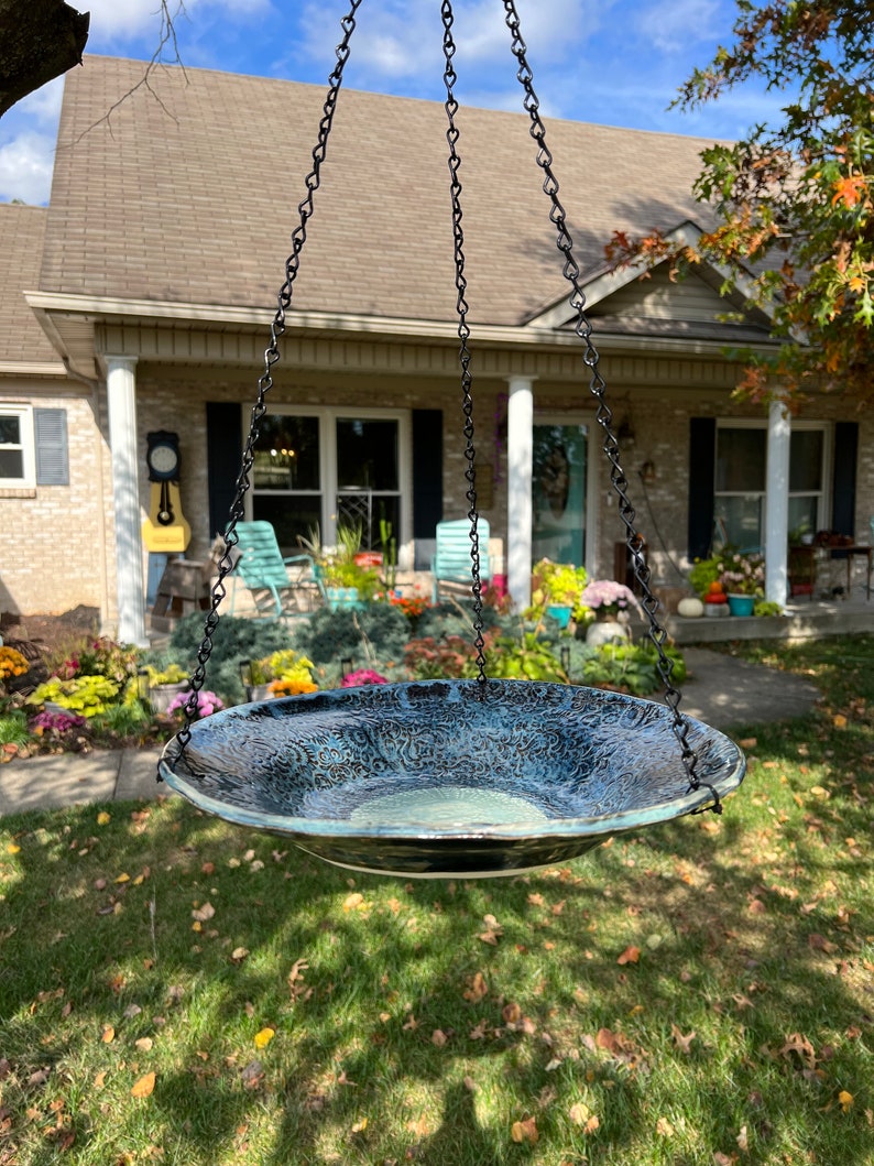 May include: A round, shallow ceramic bird bath with a textured, dark blue and black glaze featuring intricate, swirling patterns. The basin is suspended by three black metal chains attached to the rim, creating a hanging structure for outdoor use.