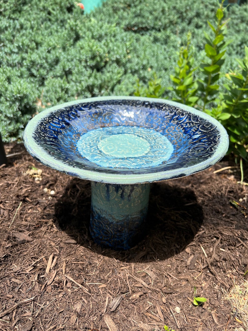 May include: A blue and green ceramic bird bath with a decorative design. The bird bath has a wide, shallow bowl and a tall, cylindrical base.