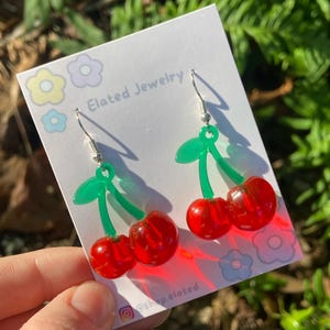 May include: A pair of cherry earrings with green stems and leaves. The earrings are made of red and green plastic and have silver hooks. The earrings are on a white card with the text "Elated Jewelry" and "@shop.elated" printed on it.