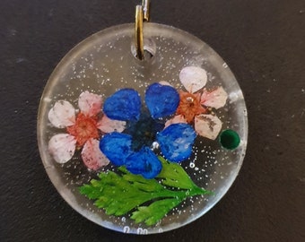 Op de afbeelding: Een ronde, transparante hars hanger met een blauwe bloem, roze en witte bloemen en groene bladeren die erin zijn geperst. De hanger heeft een gouden lus aan de bovenkant om op te hangen.