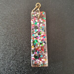 May include: A clear rectangular pendant with a gold-coloured bail. The pendant is filled with colourful glitter.
