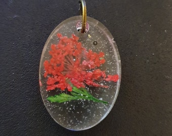 Op de afbeelding: Een heldere ovale hanger met een geperste rode bloem erin. De hanger heeft een gouden metalen lus.