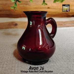 May include: A vintage ruby red cruet decanter with a handle. The glass has a glossy finish and a dark red color. The text "Avon 26" and "Vintage Ruby Red Cruet Decanter" are printed on the bottom of the image. The decanter is a decorative item.