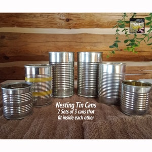 May include: A collection of six nesting tin cans, arranged on a brown surface. The cans are silver with ridged sides, and some have yellow bands. Text on the image reads "Nesting Tin Cans, 2 Sets of 3 cans that fit inside each other."