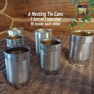 May include: Six nesting tin cans, each with a silver exterior and a gold interior. The cans are arranged in a group, with the text "6 Nesting Tin Cans" and "3 Sets of 2 cans that fit inside each other" visible.