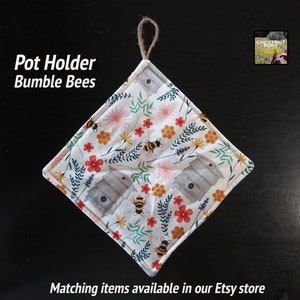 May include: A white pot holder with a bumble bee and flower design. The pot holder is square and has a loop for hanging. The text "Pot Holder Bumble Bees" is printed on the top of the image.