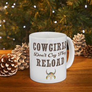 Puede incluir: Taza de cerámica blanca con el texto "COWGIRLS Don't Cry They RELOAD" en letras marrones. Una calavera decorativa con adornos florales y plumas está debajo del texto. La taza está sobre una superficie de madera.