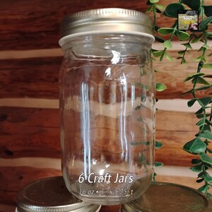 May include: Clear glass craft jar with a silver lid. The jar is labeled "6 Craft Jars" and is 16 oz, 3" x 5.25"t. The background is a wooden surface with green foliage.