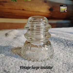 May include: A clear glass vintage surge insulator with a rounded top and a flared base. The text "SURGE" and "M.REG. PAT. PER." is embossed on the base. The text "Vintage Surge Insulator" is at the bottom of the image.