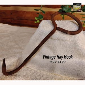 May include: A vintage hay hook made of dark brown metal. The hook is 10.75 inches long and 4.25 inches wide. The hook has a curved end and a handle. The text "Vintage Hay Hook" is printed on the image.