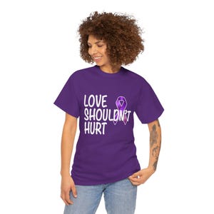 Puede incluir: Una camiseta morada con el texto blanco "LOVE SHOULDN'T HURT" y un lazo morado. La camiseta es de color liso y tiene mangas cortas.