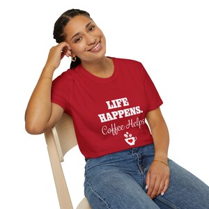 Peut inclure: Une femme portant un t-shirt rouge avec le texte "Life Happens. Coffee Helps" et un graphique de tasse de café. Elle est assise sur une chaise en bois et porte un jean bleu.