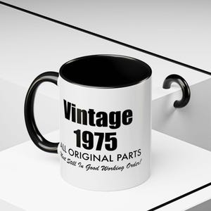 Peut inclure: Une tasse en céramique blanche avec un intérieur et une anse noirs. La tasse porte l'inscription "Vintage 1975" en lettres noires grasses, ainsi que les phrases "All Original Parts" et "Most Still In Good Working Order!"