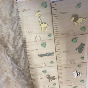 Peut inclure: Un tableau de croissance en bois sur le thème de la jungle. Le tableau présente une girafe, un crocodile, un singe, un rhinocéros, un lion, un zèbre et un éléphant. Le tableau est marqué en centimètres de 50 à 180.
