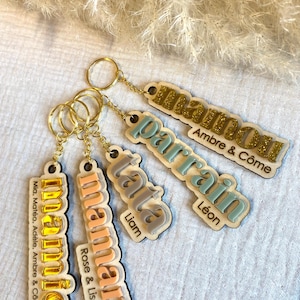 Puede incluir: Cuatro llaveros de madera con diferentes nombres y colores. Los llaveros están unidos a anillos dorados. Los nombres en los llaveros son "mamie", "maman", "tata" y "parrain".