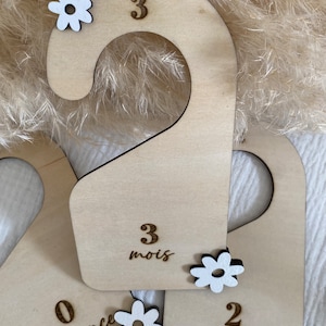 Peut inclure: Trois cintres en bois avec des embellissements de fleurs blanches. Les cintres sont étiquetés avec les chiffres 0, 2 et 3. Le cintre étiqueté 0 a également le mot "naissance" écrit dessus. Le cintre étiqueté 2 a le mot "ans" écrit dessus. Le cintre étiqueté 3 a le mot "mois" écrit dessus.