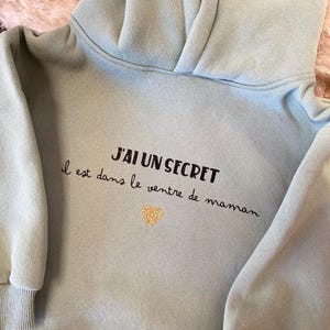 May include: A light blue hooded sweatshirt with the text "J'AI UN SECRET il est dans le ventre de maman" and a gold glitter heart.