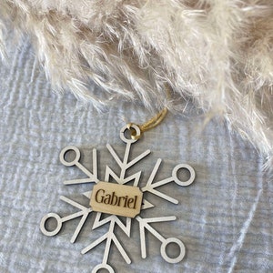 Peut inclure: Une décoration de Noël en forme de flocon de neige en bois blanc avec une ficelle dorée et une étiquette en bois sur laquelle est inscrit "Gabriel".