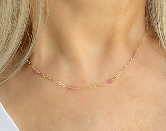 May include: A delicate gold necklace with small pink beads evenly spaced along the chain. The necklace is worn around the neck, showcasing its simple elegance. The background is a white shirt.
