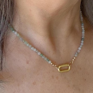 May include: A delicate necklace featuring small, light green and white beads. The necklace is secured with a gold-coloured, rectangular clasp. The beads appear to be natural stone, creating a subtle, elegant accessory.