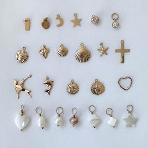 May include: A selection of gold-toned charms in diverse forms, such as a number one, a crescent, a star, a heart, a flower, a leaf, a seashell, a starfish, a cross, a bird, a dolphin, a sun, a compass rose, and a heart. Some charms have gemstones or pearls.