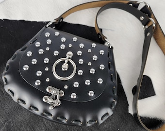 May include: Black leather shoulder bag with silver-colored studs and a decorative clasp. The bag features a stitched design around the edges and a silver ring detail. The strap is black leather with a tan underside.