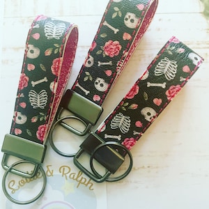 May include: Three black and white keychains with a skull and crossbones design, pink roses, and a red stitch around the edges. The keychains have black metal key rings.